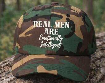 Könnte beinhalten: Eine Baseballkappe mit grünem, braunem und schwarzem Tarnmuster. Auf der Vorderseite befindet sich ein weißer Schriftzug "REAL MEN ARE Emotionally Intelligent" in einer Kombination aus fetten Blockbuchstaben und einer kursiven Schriftart.