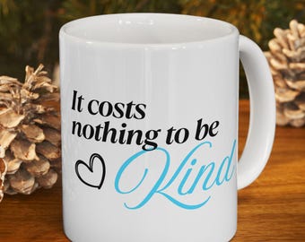 Puede incluir: Taza de cerámica blanca con el texto "It costs nothing to be Kind" en escritura negra y azul. Un corazón negro está al lado de la palabra "Kind". La taza está sobre una superficie de madera con piñas de pino en el fondo.