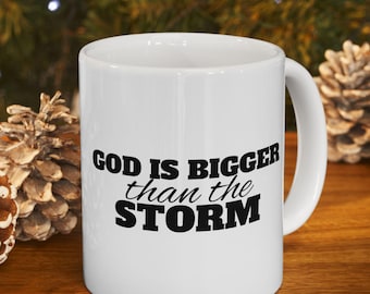 Könnte beinhalten: Weiße Keramik-Tasse mit dem Text "GOD IS BIGGER than the STORM" in Schwarz. Die Tasse steht auf einer Holzoberfläche mit Tannenzapfen und einem verschwommenen grünen Hintergrund.