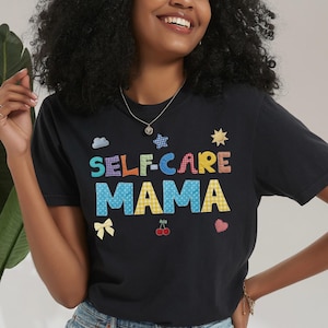 May include: Black t-shirt with the words "SELF-CARE MAMA" in colorful, patterned letters. The design includes a cloud, star, bow, cherries, and heart. The shirt is paired with denim shorts.