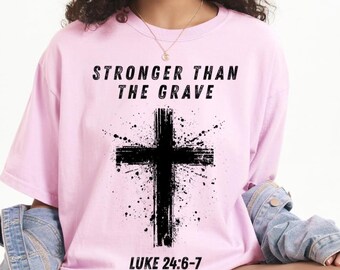 Könnte beinhalten: Ein hellrosa T-Shirt mit dem Text "STRONGER THAN THE GRAVE" über einem schwarzen Kreuz-Design. Der Text "LUKE 24:6-7" ist darunter gedruckt. Das Shirt besteht aus einem weichen Material.
