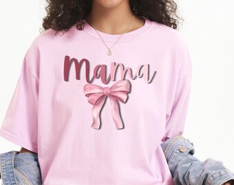 Könnte beinhalten: Hellrosa T-Shirt mit dem Wort "Mama" in dunkelrosa Schrift und einer rosa Schleifen-Grafik. Das Shirt hat eine lockere Passform und kurze Ärmel. Das Model trägt eine Jeansjacke und Shorts.