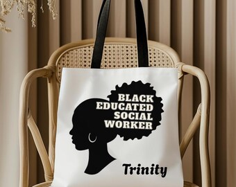 Könnte beinhalten: Weiße Tragetasche mit schwarzen Henkeln. Sie zeigt eine schwarze Silhouette einer Frau mit dem Text "BLACK EDUCATED SOCIAL WORKER" und dem Namen "Trinity". Die Tasche steht auf einem Korbstuhl.