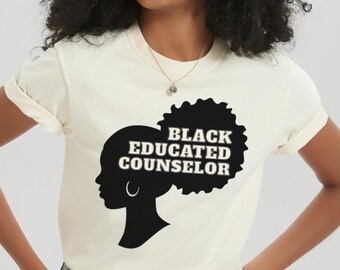 Könnte beinhalten: Cremefarbenes T-Shirt mit schwarzer Silhouette eines Frauenprofils, Afro und den Worten "BLACK EDUCATED COUNSELOR" in fetter, schwarzer Schrift. Das Shirt wird mit Jeansshorts kombiniert.