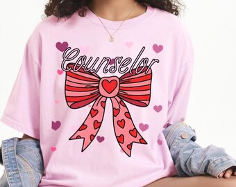 Könnte beinhalten: Hellrosa T-Shirt mit dem Wort "Counselor" in Schreibschrift über einer roten und rosa Schleife mit einem Herz in der Mitte. Die Schleife hat herzförmige Akzente. Kleine lila Herzen sind um das Design verstreut.