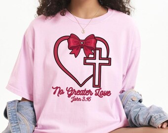 Könnte beinhalten: Hellrosa T-Shirt mit einem herzförmigen Design mit Schleife und Kreuz, mit dem Text "No Greater Love John 3:16" in einer Schriftart. Das Shirt hat eine lockere Passform.