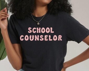 Könnte beinhalten: Schwarzes T-Shirt mit den Worten "SCHOOL COUNSELOR" in rosa, donutartigen Buchstaben. Das Model trägt Jeansshorts und weiße Turnschuhe. Eine Halskette und ein Armband sind ebenfalls zu sehen.