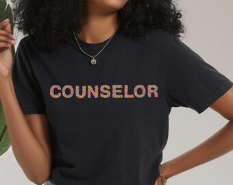 Könnte beinhalten: Schwarzes T-Shirt mit dem Wort "COUNSELOR" in bunten, strukturierten Buchstaben. Das Shirt hat kurze Ärmel und wird mit Jeansshorts getragen. Das Bild ist bei natürlichem Licht aufgenommen.