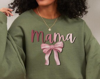 Könnte beinhalten: Olivgrünes Sweatshirt mit dem Wort "Mama" in einem rosa Farbverlauf und einer rosa Schleifen-Grafik. Das Sweatshirt hat lange Ärmel und eine lockere Passform.