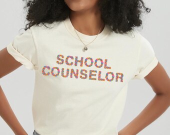 Könnte beinhalten: Cremefarbenes T-Shirt mit dem Schriftzug "SCHOOL COUNSELOR" in bunten, gesprenkelten Buchstaben. Das Shirt hat hochgekrempelte Ärmel und wird mit Jeansshorts kombiniert. Eine Halskette und Armbänder sind ebenfalls zu sehen.