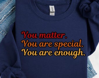 Puede incluir: Sudadera azul marino con el texto "You matter. You are special. You are enough." en rojo, naranja y amarillo. La sudadera está doblada sobre unos vaqueros azules.