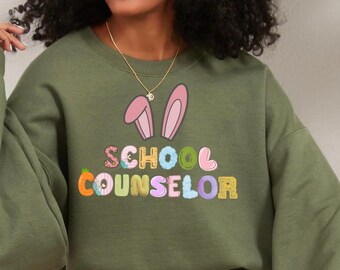 Könnte beinhalten: Ein olivgrünes Sweatshirt mit dem Schriftzug "SCHOOL COUNSELOR" in bunten, verspielten Buchstaben. Das Design umfasst Hasenohren und eine Karotte und sorgt für einen lustigen, frühlingshaften Look.