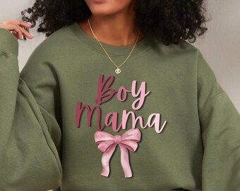 Könnte beinhalten: Ein olivgrünes Sweatshirt mit Rundhalsausschnitt, das den Schriftzug "Boy Mama" in einer rosafarbenen Verlaufsschrift über einer hellrosa illustrierten Schleifengrafik zeigt. Das Sweatshirt hat lange Ärmel und einen gerippten Ausschnitt.