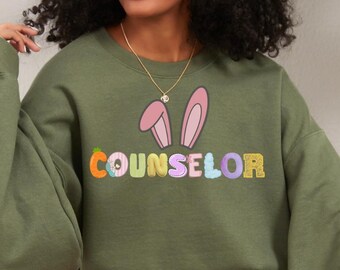 Könnte beinhalten: Ein olivgrünes Sweatshirt mit dem Wort "COUNSELOR" in bunten, gestickten Buchstaben. Über dem Wort befindet sich eine Grafik von rosa Hasenohren. Das Sweatshirt ist ein lässiges Kleidungsstück mit langen Ärmeln.