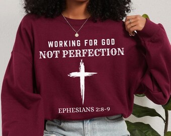 Könnte beinhalten: Ein weinrotes Sweatshirt mit dem Text "WORKING FOR GOD NOT PERFECTION" und einer weißen Kreuzgrafik. Der Text "EPHESIANS 2:8-9" ist darunter gedruckt. Das Sweatshirt hat einen lässigen Stil.