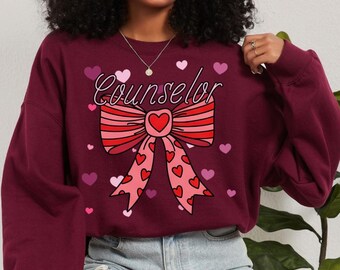Könnte beinhalten: Burgunderfarbenes Sweatshirt mit dem Wort "Counselor" in Schreibschrift über einem großen Schleifendesign. Die Schleife ist rot und rosa mit Herzakzenten. Kleine rosa und rote Herzen sind um das Design verstreut.