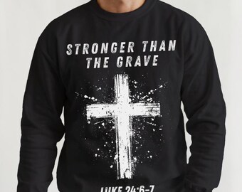 Könnte beinhalten: Schwarzes Sweatshirt mit weißem Kreuz-Design und dem Text "STRONGER THAN THE GRAVE" und "LUKE 24:6-7". Das Kreuz hat einen Used-Look mit Spritzereffekt. Das Sweatshirt ist ein Rundhals-Stil.