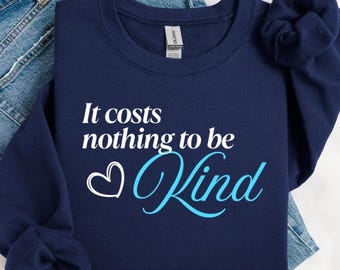 Puede incluir: Una sudadera azul marino con el texto "It costs nothing to be Kind" en blanco y azul claro. Un corazón blanco está al lado de la palabra "Kind". La sudadera está doblada sobre unos vaqueros azules.