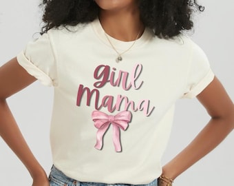Könnte beinhalten: Cremefarbenes T-Shirt mit den Worten "Girl Mama" in rosa Schrift und einer rosa Schleifen-Grafik. Das Shirt hat kurze Ärmel und ist aus weichem Material. Das Model trägt blaue Jeansshorts.