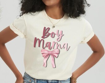 Könnte beinhalten: Cremefarbenes T-Shirt mit dem Text "Boy Mama" in rosa Schrift und einer rosa Schleifen-Grafik. Das Shirt hat hochgekrempelte Ärmel und ist aus weichem Material.