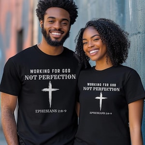 May include: Two people wearing black t-shirts with the text "WORKING FOR GOD NOT PERFECTION" and a cross graphic. The text "EPHESIANS 2:8-9" is below the cross. The people are smiling.