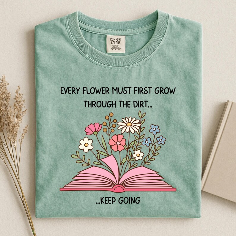 Puede incluir: Camiseta verde claro con el texto "EVERY FLOWER MUST FIRST GROW THROUGH THE DIRT... ...KEEP GOING". La camiseta presenta un gr&aacute;fico de un libro rosa abierto con flores coloridas que brotan de &eacute;l. La camiseta est&aacute; doblada.