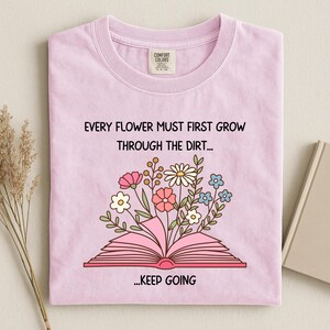Puede incluir: Camiseta de color lila claro con el texto "EVERY FLOWER MUST FIRST GROW THROUGH THE DIRT... ...KEEP GOING". La camiseta presenta un gr&aacute;fico de un libro abierto con flores coloridas que brotan de &eacute;l. Una etiqueta Comfort Colors es visible.