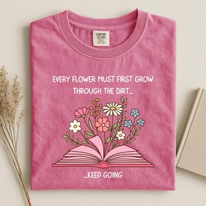 Puede incluir: Camiseta rosa con la frase "EVERY FLOWER MUST FIRST GROW THROUGH THE DIRT... ...KEEP GOING". Una ilustraci&oacute;n floral colorida emerge de un libro abierto. La camiseta tiene cuello redondo y una etiqueta.