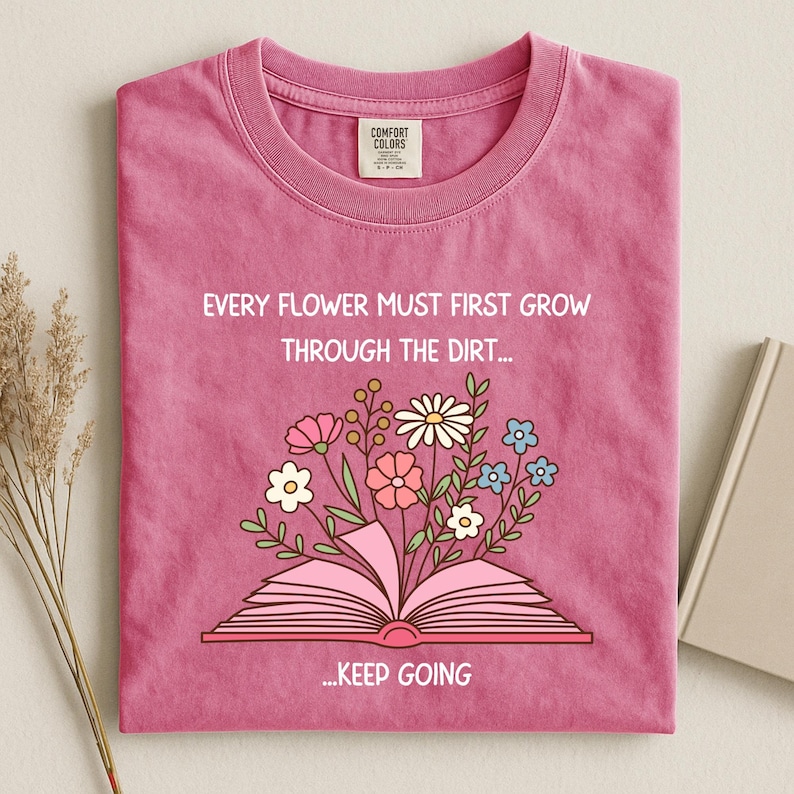 Puede incluir: Camiseta rosa con la frase "EVERY FLOWER MUST FIRST GROW THROUGH THE DIRT... ...KEEP GOING". Una ilustraci&oacute;n floral colorida emerge de un libro abierto. La camiseta tiene cuello redondo y una etiqueta.