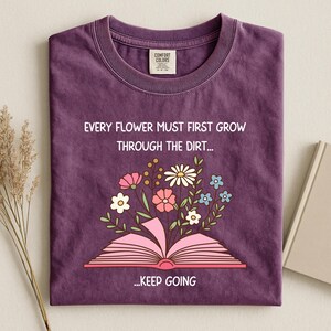 Puede incluir: Una camiseta morada con el texto "EVERY FLOWER MUST FIRST GROW THROUGH THE DIRT... ...KEEP GOING". La camiseta presenta un dise&ntilde;o floral con un libro abierto y varias flores rosas, blancas y azules.