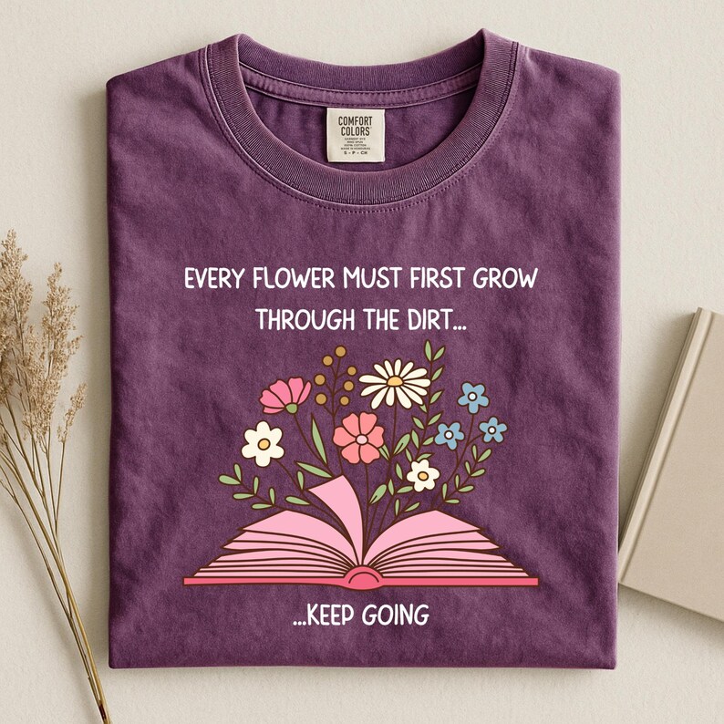 Puede incluir: Una camiseta morada con el texto "EVERY FLOWER MUST FIRST GROW THROUGH THE DIRT... ...KEEP GOING". La camiseta presenta un dise&ntilde;o floral con un libro abierto y varias flores rosas, blancas y azules.