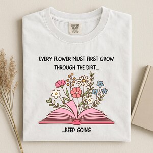 Puede incluir: Camiseta blanca con cuello redondo con un dise&ntilde;o floral que emerge de un libro rosa abierto. El texto dice: "EVERY FLOWER MUST FIRST GROW THROUGH THE DIRT... ...KEEP GOING."