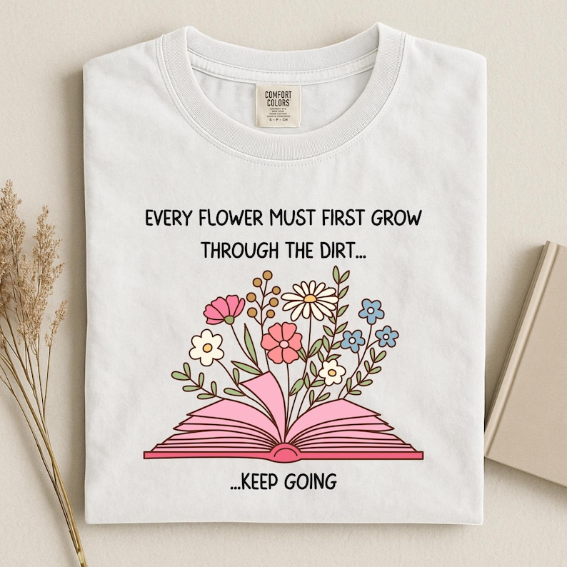 Puede incluir: Camiseta blanca con cuello redondo con un dise&ntilde;o floral que emerge de un libro rosa abierto. El texto dice: "EVERY FLOWER MUST FIRST GROW THROUGH THE DIRT... ...KEEP GOING."