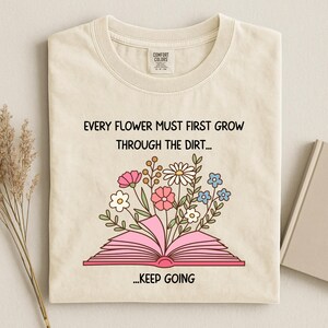 Puede incluir: Camiseta color crema con la cita "EVERY FLOWER MUST FIRST GROW THROUGH THE DIRT... ...KEEP GOING" y un gr&aacute;fico de un libro rosa abierto con flores coloridas. La camiseta tiene cuello redondo y una etiqueta.