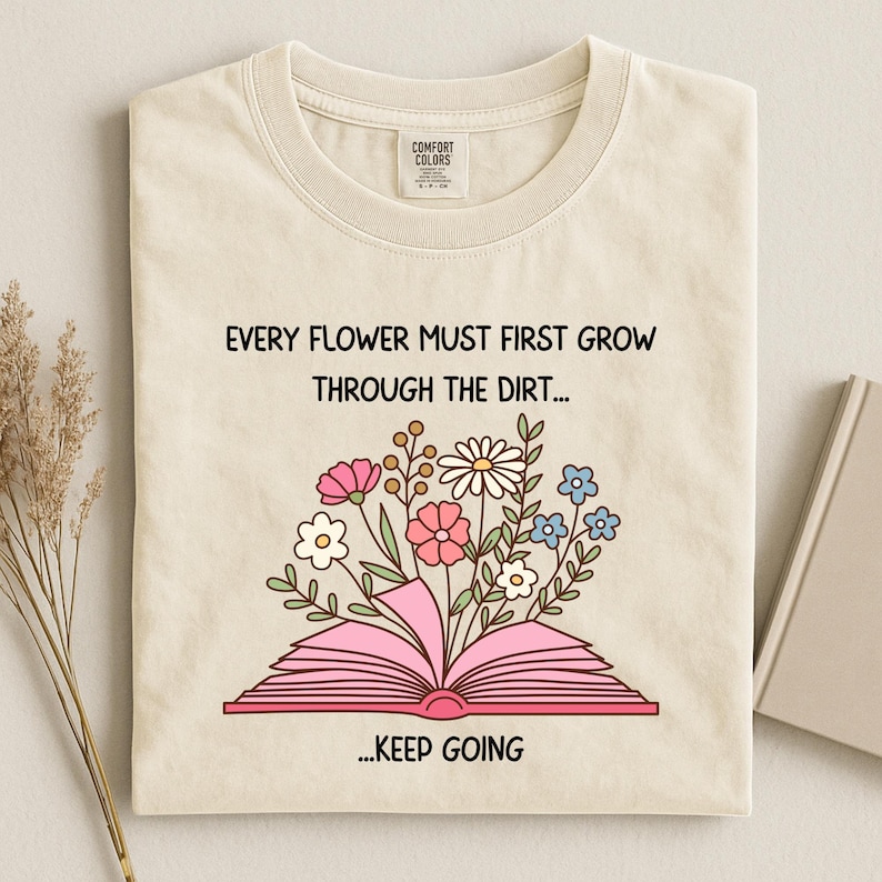 Puede incluir: Camiseta color crema con la cita "EVERY FLOWER MUST FIRST GROW THROUGH THE DIRT... ...KEEP GOING" y un gr&aacute;fico de un libro rosa abierto con flores coloridas. La camiseta tiene cuello redondo y una etiqueta.