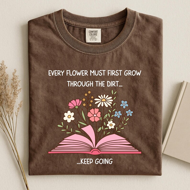 Puede incluir: Camiseta marr&oacute;n con el texto "EVERY FLOWER MUST FIRST GROW THROUGH THE DIRT... ...KEEP GOING". La camiseta presenta un gr&aacute;fico de un libro rosa abierto con flores coloridas que brotan de &eacute;l. La camiseta est&aacute; doblada.