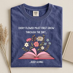 Puede incluir: Una camiseta azul marino con el texto "EVERY FLOWER MUST FIRST GROW THROUGH THE DIRT... ...KEEP GOING." La camiseta presenta un gr&aacute;fico de un libro rosa abierto con flores coloridas que brotan de &eacute;l.