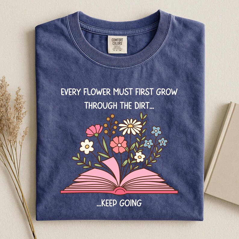 Puede incluir: Una camiseta azul marino con el texto "EVERY FLOWER MUST FIRST GROW THROUGH THE DIRT... ...KEEP GOING." La camiseta presenta un gr&aacute;fico de un libro rosa abierto con flores coloridas que brotan de &eacute;l.