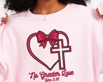 Könnte beinhalten: Hellrosa Sweatshirt mit einem Herz- und Kreuz-Design. Eine rote Schleife sitzt auf dem Herzen, darunter die Worte "No Greater Love" und "John 3:16" in einer Schriftart. Das Sweatshirt hat eine einfarbige hellrosa Farbe.
