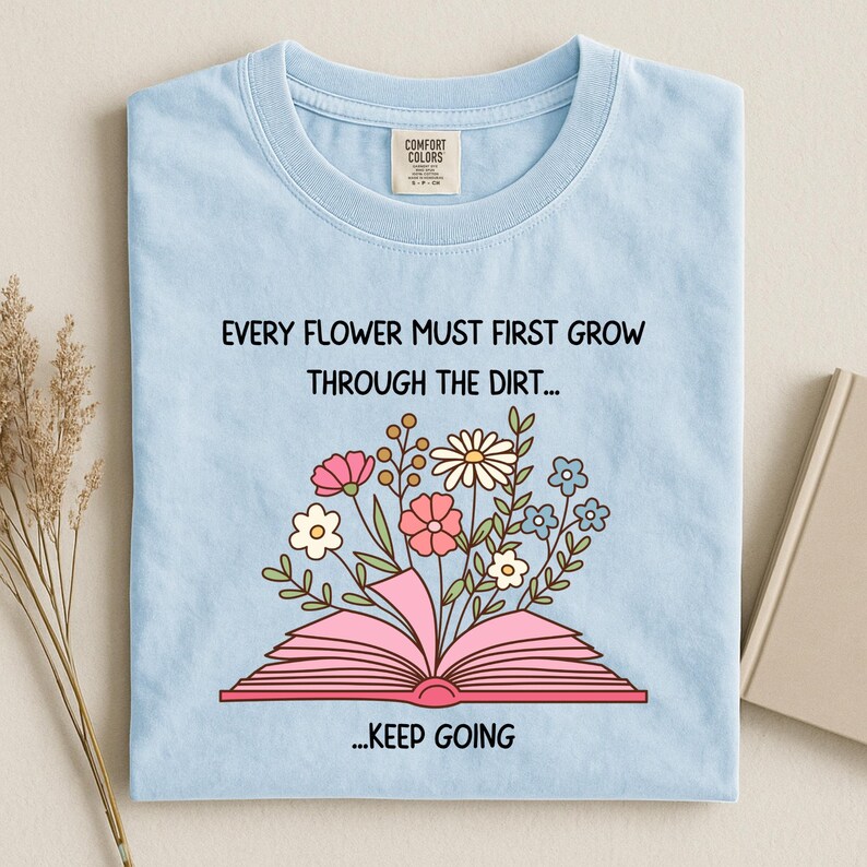 Puede incluir: Camiseta azul claro con el texto "EVERY FLOWER MUST FIRST GROW THROUGH THE DIRT... ...KEEP GOING." La camiseta presenta un gr&aacute;fico de un libro rosa abierto con flores coloridas que florecen.