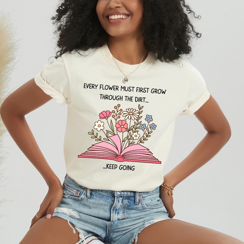 Puede incluir: Camiseta color crema con el texto "EVERY FLOWER MUST FIRST GROW THROUGH THE DIRT..." sobre una ilustraci&oacute;n de un libro abierto con flores. Debajo est&aacute; el texto "...KEEP GOING".