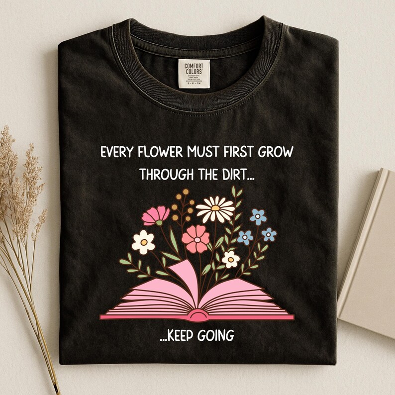 Puede incluir: Camiseta negra con la cita "EVERY FLOWER MUST FIRST GROW THROUGH THE DIRT... ...KEEP GOING" en blanco. La camiseta presenta un libro rosa abierto con flores coloridas. Las flores incluyen rosa, blanco y azul.