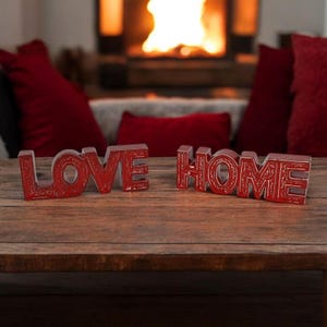 Pode incluir: Sinais decorativos vermelhos de madeira com as palavras "LOVE HOME" numa mesa de madeira. As letras são esculpidas com um padrão decorativo. O fundo apresenta uma lareira e almofadas vermelhas, criando uma atmosfera acolhedora e quente.