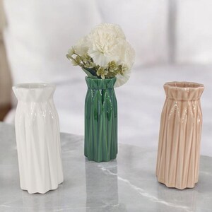 Pode incluir: Três vasos de cerâmica em branco, verde e pêssego. O vaso branco tem um design em espiral, o vaso verde contém flores brancas e o vaso pêssego tem ranhuras verticais. Estes vasos decorativos são adequados para decoração de casa.