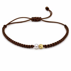 May include: Brown braided bracelet featuring a white pearl and a gold-colored bead. The bracelet has an adjustable sliding knot closure. The jewelry is displayed against a white background.