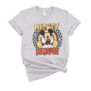 May include: A light gray t-shirt featuring a retro-style graphic of Mickey Mouse. The design includes the words "Mickey Mouse" in a colorful, circular design with stars and a checkered pattern. The shirt has short sleeves and a crew neck.
