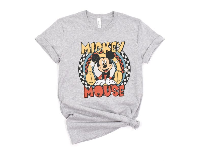 May include: A light gray t-shirt featuring a retro-style graphic of Mickey Mouse. The design includes the words "Mickey Mouse" in a colorful, circular design with stars and a checkered pattern. The shirt has short sleeves and a crew neck.