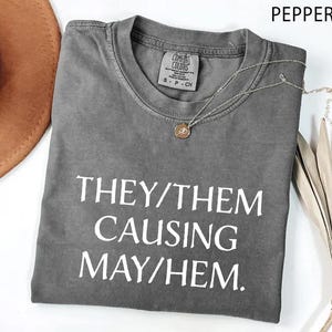 May include: A gray t-shirt with the text "THEY/THEM CAUSING MAYHEM" in white. The shirt is folded and has a round neckline. A gold-colored necklace and a brown hat are next to the shirt.