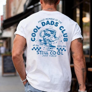 May include: White t-shirt with a blue graphic on the back. The graphic features a coffee mug riding a skateboard, with the text "COOL DADS CLUB" and "STILL COOL JUST TIRED". The shirt is a casual style.