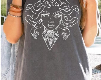 May include: Dark gray t-shirt featuring a white graphic of Medusa's face with snakes for hair. The design includes a floral element at the neck. The shirt is a casual style, suitable for everyday wear.